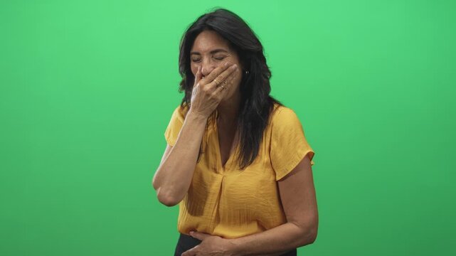 Woman covers mouth with hand and clasps stomach in studio wearing yellow blouse, eyes closed and smiling into her palm; embarrassment vulnerability.