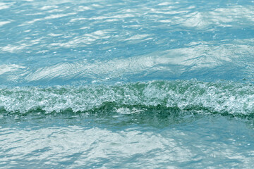 Naklejka premium A close-up view of a small turquoise sea wave gently breaking on a calm water surface, showing ripples, texture and sparkling droplets that evoke freshness, tranquility and coastal summer mood.
