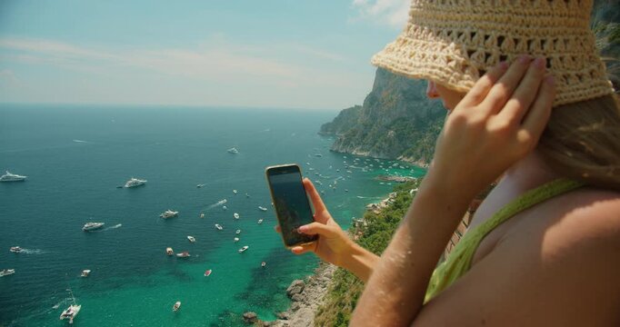 Young woman recording phone video of boats on the blue sea from a sunny viewpoint in Capri