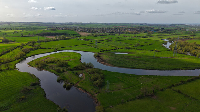 Aerial view of the River Trent winding through lush green meadows and farmland with electricity pylons under a cloudy sky Repton, England, United Kingdom.