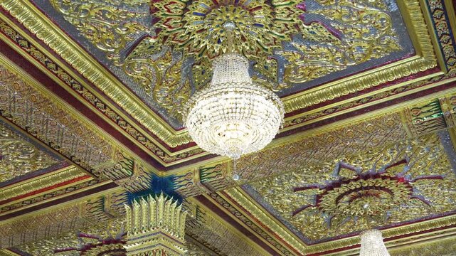 A crystal chandelier hangs beneath an ornate temple ceiling filled with gold patterns and mirrored details. The interior view highlights luxury, symmetry, and rich decorative craft.