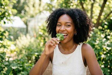 Fototapeta premium Happy young black woman happily eating a fresh broccoli floret in a green garden