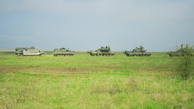 A wide shot showing a line of heavy military tanks and armored personnel carriers moving in a convoy through a green grassy field under an overcast sky during a deployment or training exercise