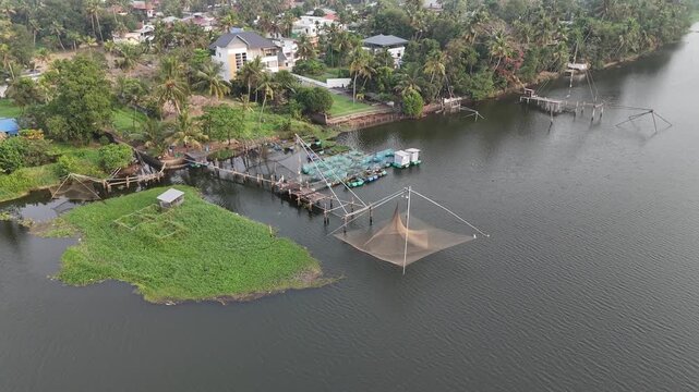 Drone footage captures traditional Chinese fishing nets suspended over calm backwaters alongside floating fish farm cages. Lush green vegetation and palm trees line the waterfront with residential bui
