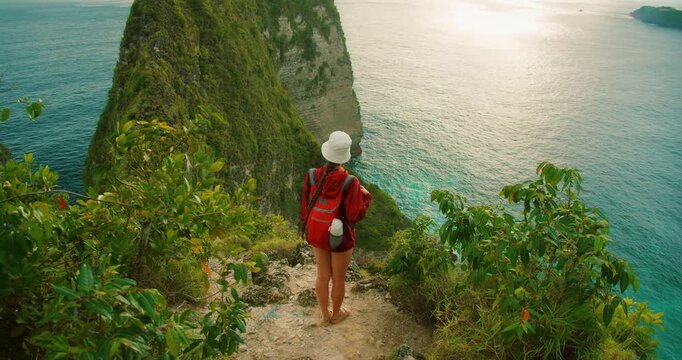 Woman with backpack looks at steep green cliff and blue ocean at Kelingking Nusa Penida Bali
