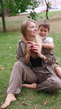Vertical video. Family outdoor. Parent love. Smiling happy mother on green grass enjoying son hugging wrapping arms tenderly around neck in park.