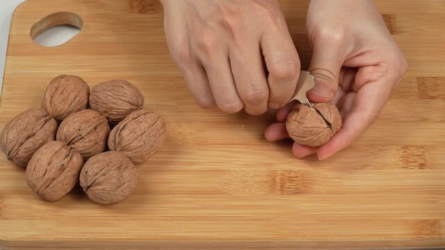 Close-up of female hands opening a whole walnut with a metal cracking tool on a wooden board. Authentic healthy kitchen food preparation concept.