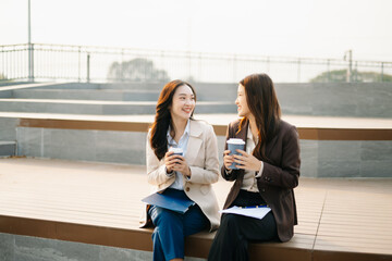 Fototapeta premium Asian businesswomen reviewing documents and writing notes outdoors. Modern teamwork, planning, and professional collaboration in a casual corporate lifestyle setting.