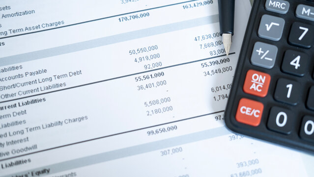 Pen pointing to financial figures next to calculator on report paper, accounting, liability review and business finance concept with office desk background.