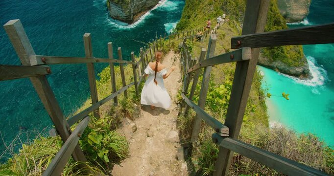 Woman in white dress walks down steep path at Kelingking Beach in Nusa Penida