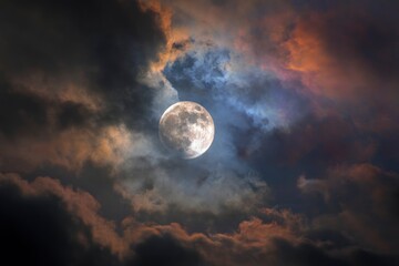 Obraz premium Bright full moon glowing behind dramatic dark storm clouds, ethereal night sky with colorful orange and blue atmosphere.
