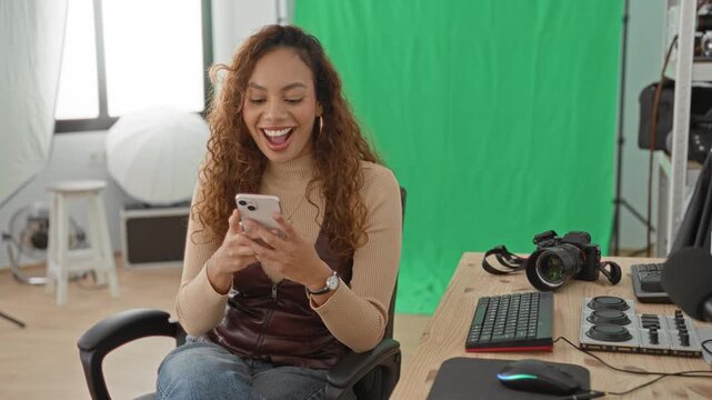 Woman holding smartphone smiles and raises hand in studio with green screen and camera gear; joyful celebration.