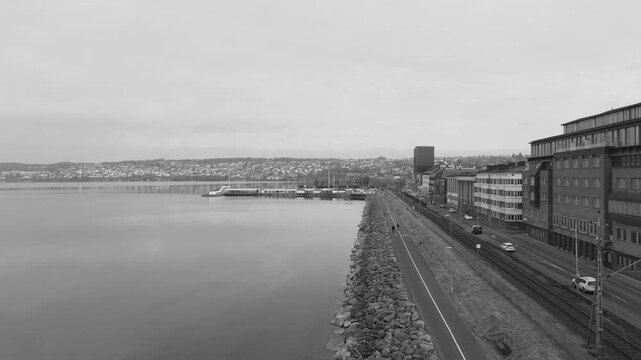 Black and white drone video capturing the serene Lake Zurich waterfront in Horgen, Switzerland. Aerial footage reveals the tranquil lake alongside a stone-lined promenade with urban views, no people.