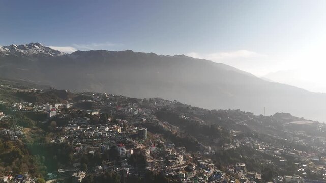 Drone footage reveals a sprawling hill station town built on steep mountain slopes with densely packed buildings and houses. Snow-capped Himalayan peaks rise majestically in the background under a haz