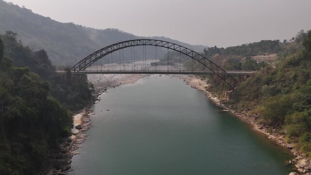 Drone footage reveals a steel arch bridge spanning a calm turquoise river flowing through lush green forested hills. Rocky riverbanks and hazy mountains create a serene landscape in northeastern India