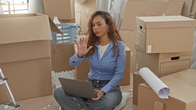 Woman pointing hand at stacked cardboard moving boxes beside a ladder and packing materials while seated with laptop amid relocation in a building; focused planning.