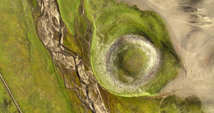Top-down drone shot of a green volcanic crater beside braided runoff channels and pale ash terrain, revealing abstract natural patterns in a remote highland landscape.