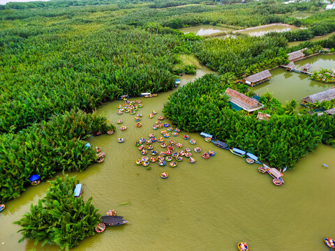 Aerial view of traditional round basket boats navigating a river through the lush Bay Mau coconut forest with thatched roof huts in Hoi An, An Giang, Vietnam.