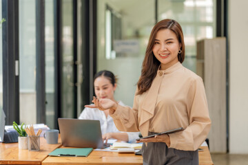 Obraz premium Smiling Asian businesswoman welcoming and gesturing to present her office teamwork with laptop and tablet.
