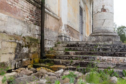 Weathered stone steps lead to a large classical column beside peeling plaster and exposed brick, with moss, weeds and rubble highlighting decay, neglect and historic architectural details
