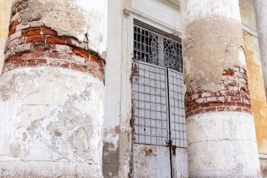 Weathered classical columns flank a rusted metal gate on a historic building, revealing exposed red brick, peeling plaster and textured decay that conveys age, neglect and architectural character