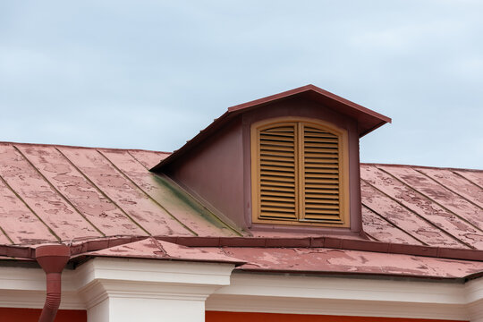 Weathered red metal roof featuring a dormer with closed wooden shutters above a white classical facade; peeling paint, gutter detail and cloudy sky convey rustic character and architectural texture