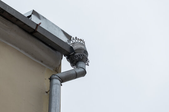 Close-up of a metal gutter and curved downspout attached to a pale building corner, highlighting roof line details, weathered metal textures and functional drainage elements against an overcast sky