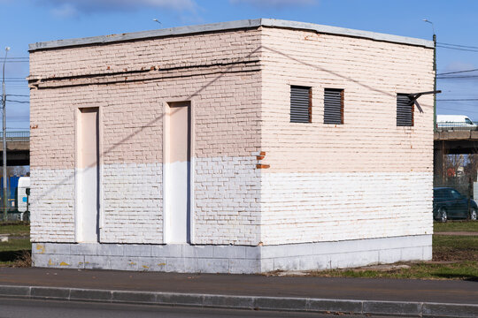 A small urban utility building with white painted brick, minimal windows and ventilation grilles, sitting beside a road and industrial overpass. Weathered facade and simple geometric architecture