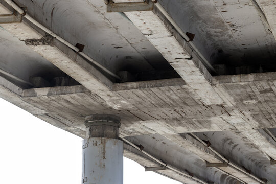 Close-up view of a weathered concrete bridge underside showing support pillar, beams and pipes; industrial overpass infrastructure conveying urban decay, texture, engineering detail and transportation