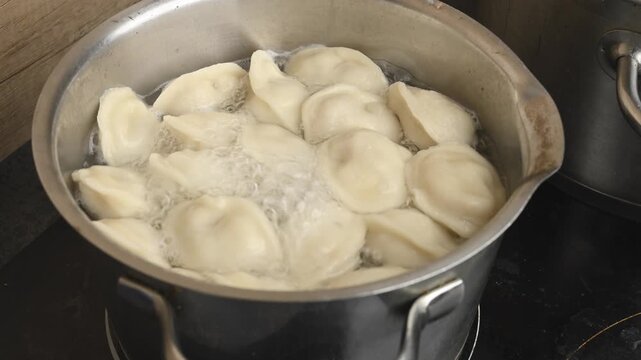 Many light-colored dumplings boil vigorously in a metal pot on a stovetop in a home kitchen. The water bubbles around them during preparation