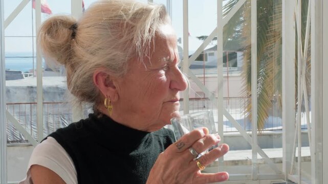 Senior blonde woman drinking a glass of water on a bright terrace. Elderly lady staying hydrated outdoors during a sunny day. Concept of healthy aging, wellness, and retirement lifestyle
