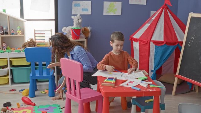 Boy drawing with crayon and paper at a small red table while a teacher leans beside him near toys and tent in a playroom building; learning calm.