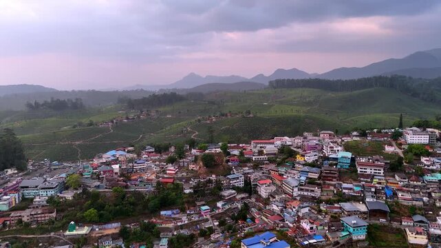Cinematic aerial view of Valparai showcasing a hillside town with colorful rooftops, surrounded by lush tea plantations and rolling hills under a dramatic evening sky with soft pink hues.