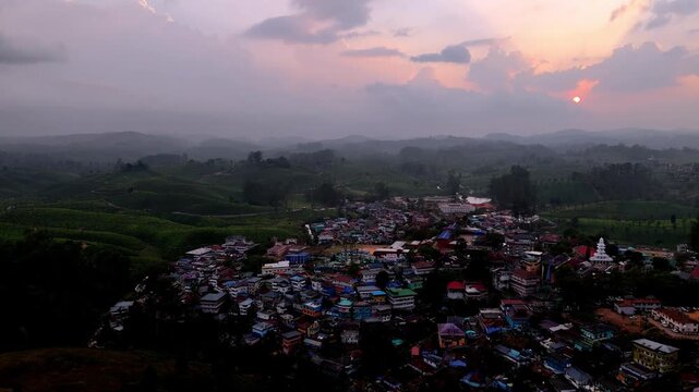 Cinematic aerial view of Valparai showcasing a compact hillside town with colorful rooftops surrounded by lush tea plantations, under a glowing sunset sky with warm tones and misty hills.