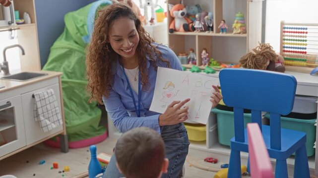 Teacher points finger to paper with abc drawing while smiling to a little boy in a preschool playroom building; learning joy.
