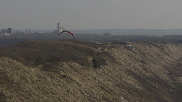 Drone pulls back showing paragliders over Zoutelande dunes with distant industry