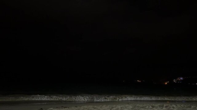 Intense lightning flash illuminating dark storm clouds over the Andaman Sea. Dramatic night scenery of Phuket beach with white ocean waves and city lights on hills, authentic monsoon photo.