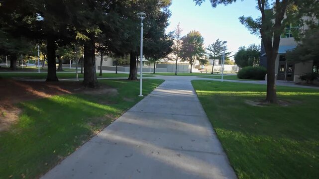 Point of view video walking along a concrete path through the lush green grounds of Modesto Junior College in California, surrounded by trees and modern buildings on a sunny day