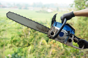 Fototapeta premium Close-up of a chainsaw held by a worker wearing gloves in a green rural field, ready for cutting wood. Concept of agriculture, forestry, maintenance, and outdoor labor work.