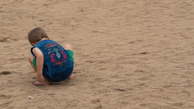 a young boy seen from behind crouching and playing on a wide sandy beach during a summer vacation, wearing a blue tank top with a tropical bali theme