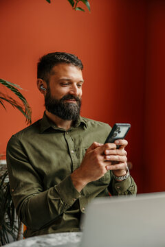 Bearded man using smartphone and earbuds while working remotely