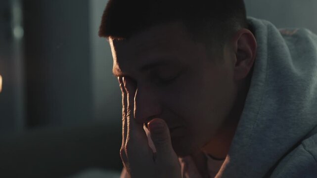 IT specialist working remotely suffering from eye strain after long hours at the computer. He removes his glasses and rubs his eyes in a dark evening room. Concept of digital workload, fatigue, and mo