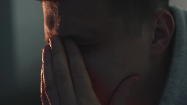 Man in grey hoodie suffering from eye strain after long hours of computer work. He removes his glasses and rubs his eyes in a dim room. Concept of vision problems, fatigue, and remote IT work stress.