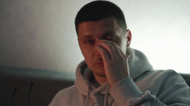 Young man experiencing eye pain and irritation after prolonged screen time. He removes his glasses and rubs his eyes in a dimly lit room. Concept of vision strain, computer fatigue, and eye health pro