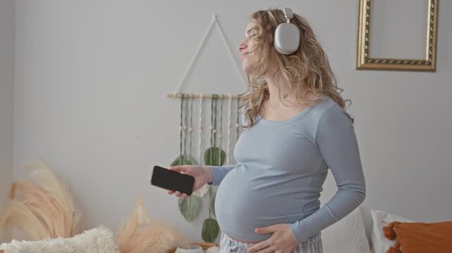 Pregnant woman wearing headphones holding smartphone with hand on belly and eyes closed smiling in a bedroom studio; serenity.