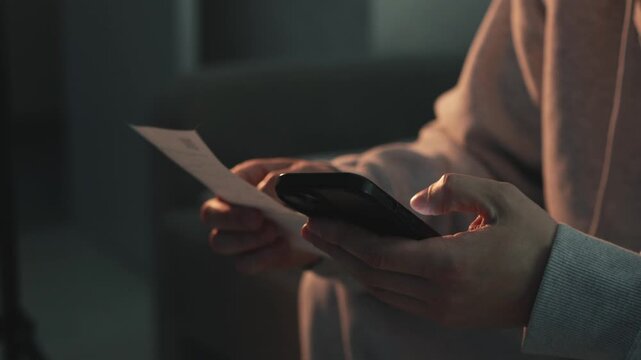 Close-up of male hands holding a paper receipt while using a smartphone in a dark room. Concept of online payment, bill processing, and financial management. Focus on modern digital transactions