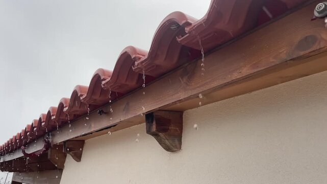 A detailed horizontal shot of a rotting wooden structural beam caused by a broken gutter. Rain pours directly onto the decaying timber beneath an old tiled roof. High quality 4k footage