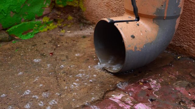 Close-up water pouring from worn gutter pipe onto stone pavement during rain, creating moody and cinematic scene. Perfect for urban texture backgrounds, drainage concepts, rainy day footage