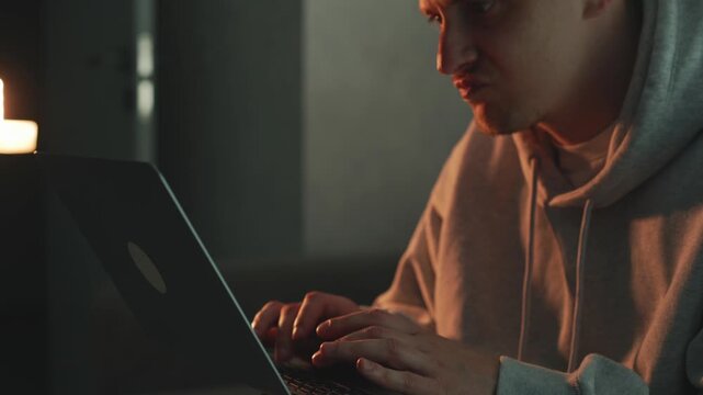 Young freelancer working on a laptop in his room in the evening. He types on the keyboard, focused on remote IT work and programming tasks. Concept of home office, digital profession, and productivity
