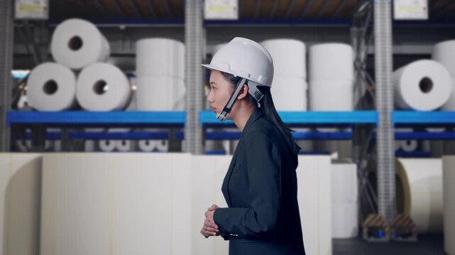 Side View of Asian Businesswoman Walking and Speech at Warehouse with Rows of Stacked White Polymer and Paper Rolls